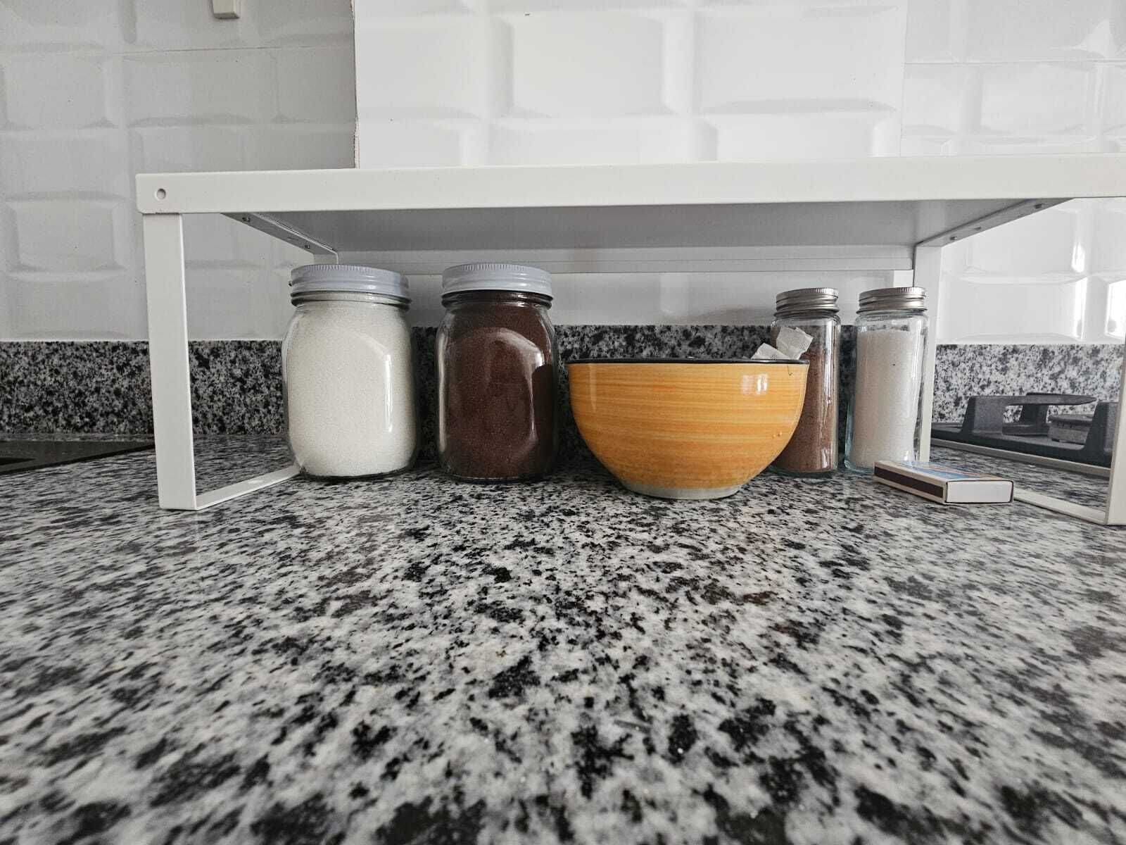 A kitchen counter with granite backsplash, white cabinets, and a white shelf holding jars and a bowl.