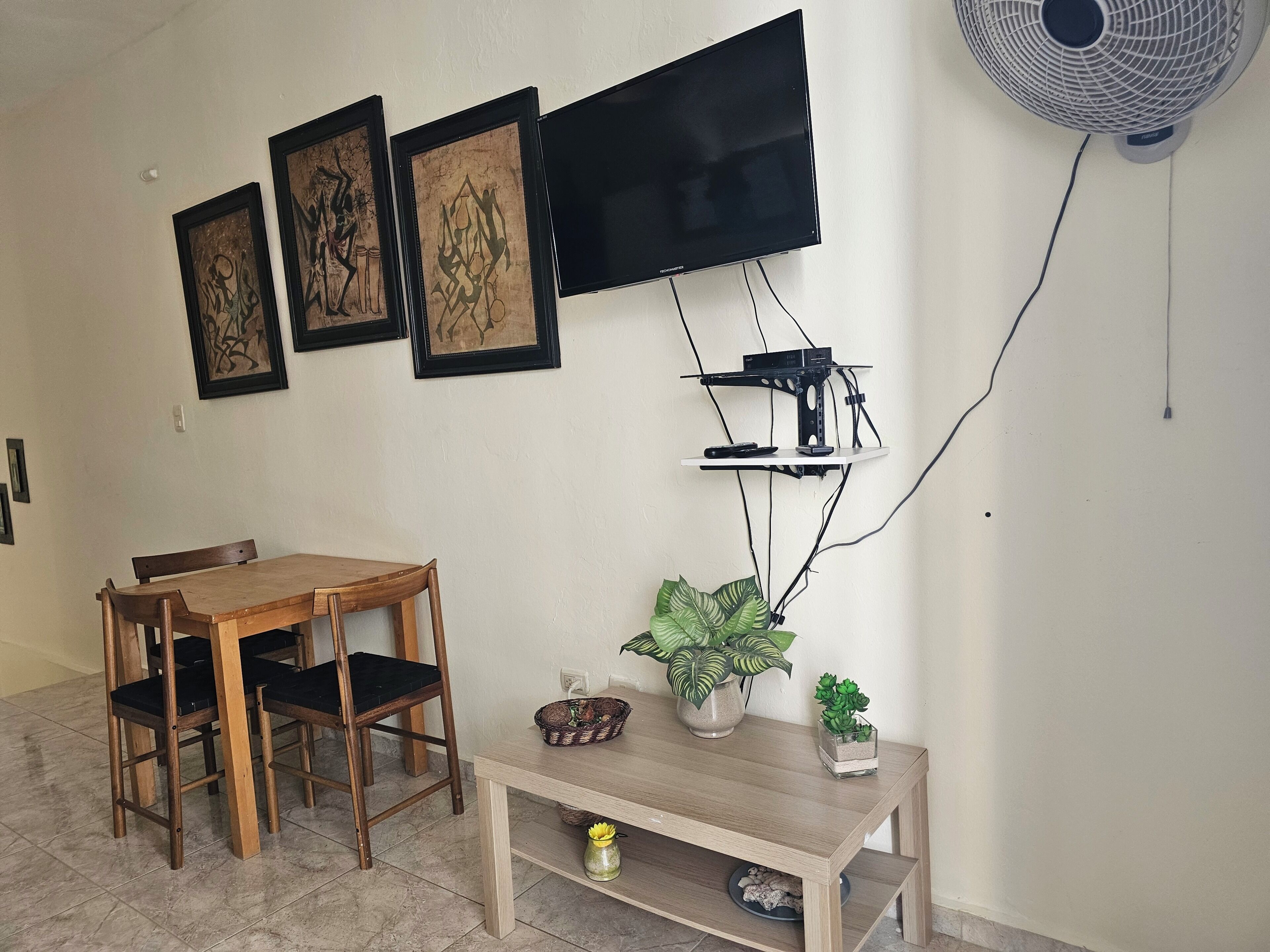 Un coin repas avec une table et des chaises en bois, un téléviseur fixé au mur et un ventilateur mural.
