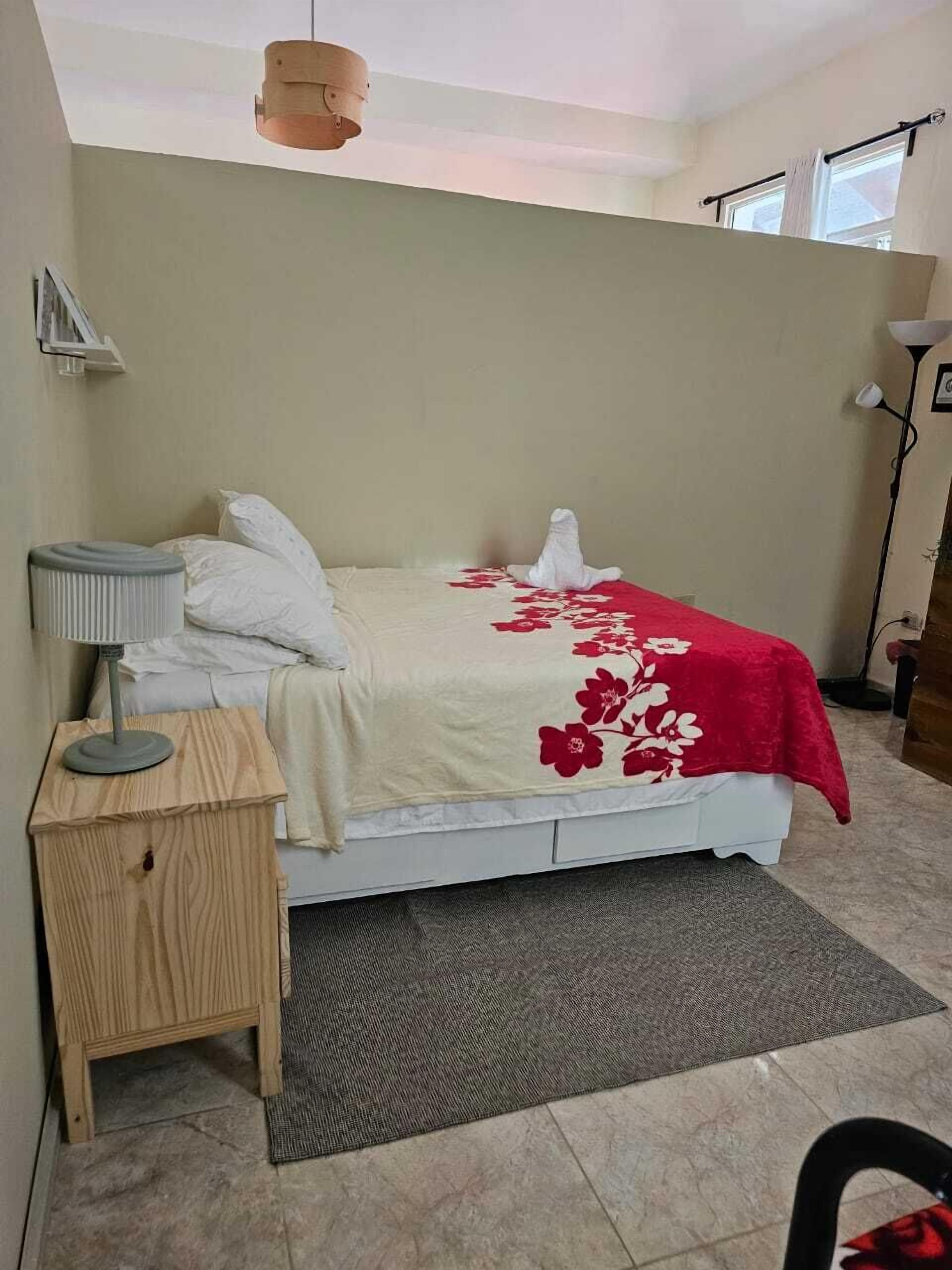 A bedroom with a bed, bedside table, lamp, and a view of the sky through a window.
