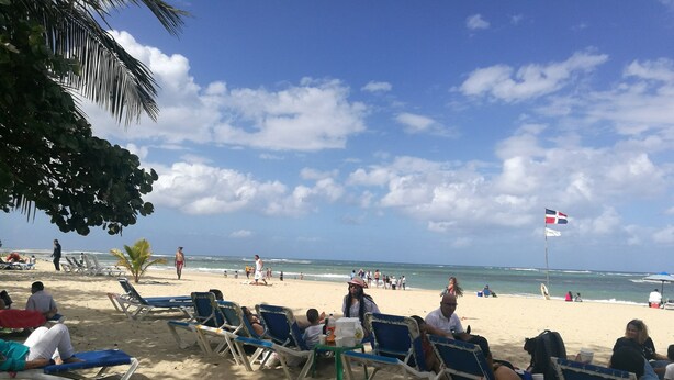 A beach with rows of blue and white lounge chairs, a flag, and people relaxing. - Apartamento Costa Azul en El Pueblito (Puerto Plata)