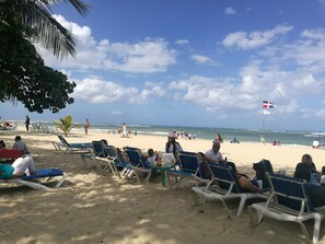 A beach with rows of blue and white lounge chairs, a flag, and people relaxing. - Apartamento Costa Azul en El Pueblito (Puerto Plata)