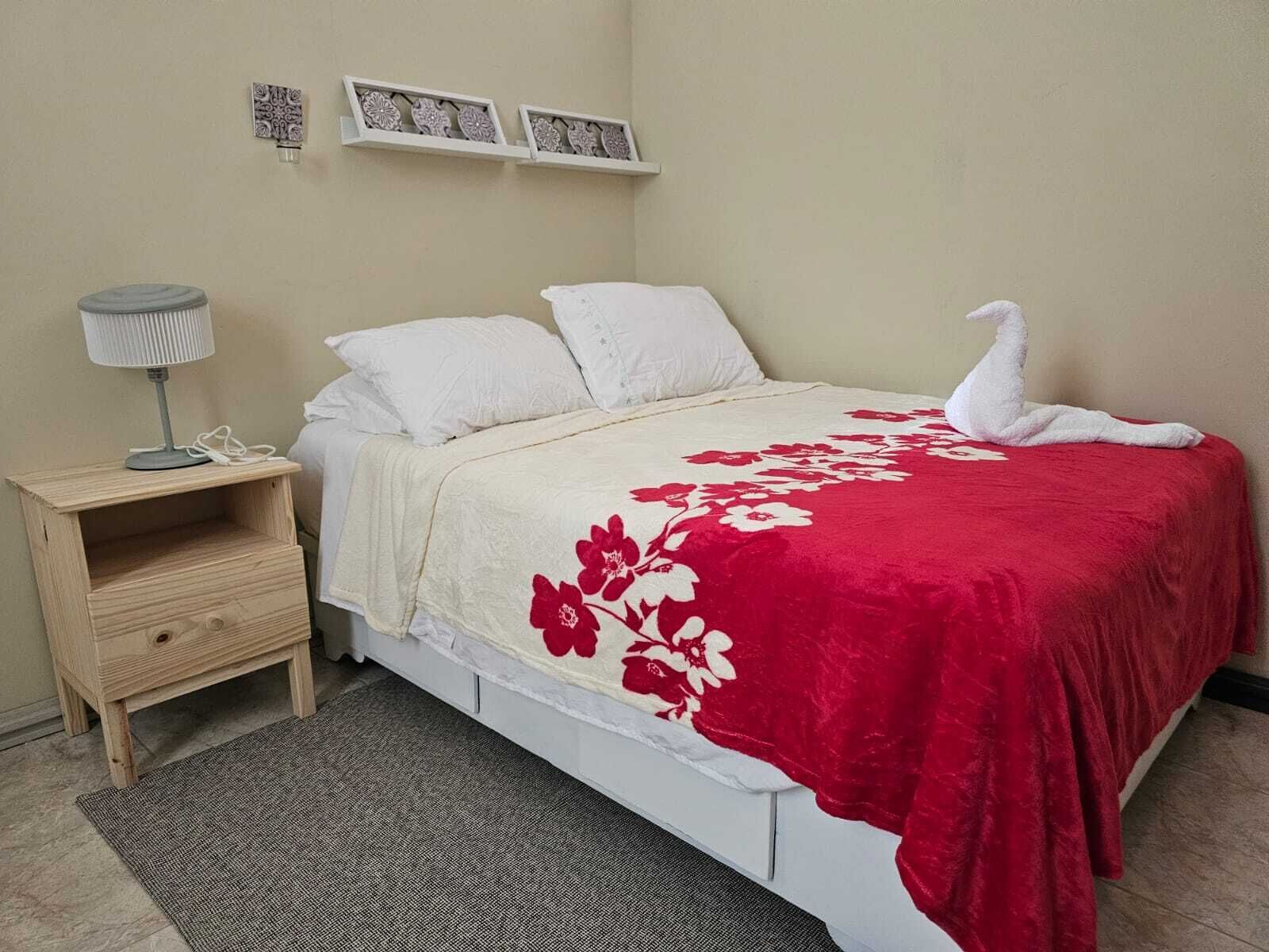 Una cama bien hecha con una colcha floral roja, almohadas blancas y una decoración de cisne blanco. Hay una mesita de noche de madera con una lámpara y una alfombra gris en el suelo.