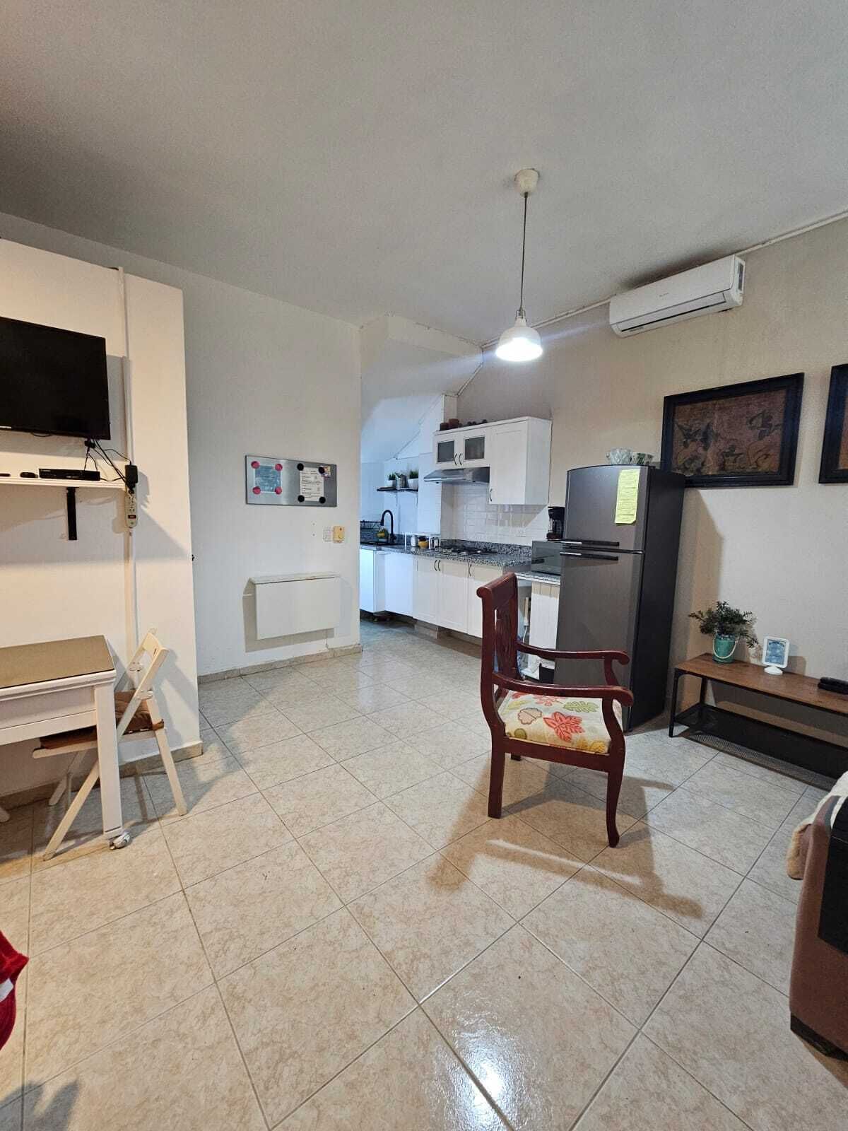 A compact kitchen with white cabinets, a wooden chair, a dining table, and a TV mounted on the wall.