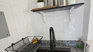 A kitchen with a granite countertop, a black sink, a dish rack, and a shelf with kitchen utensils and plants. - Apartamento Costa Azul en El Pueblito (Puerto Plata)