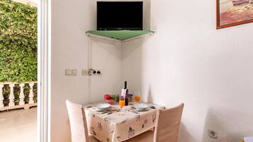 A corner room with a television mounted on a green shelf, a table set with plates, a bottle, and glasses, and a view of greenery outside.