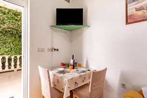 A corner room with a television mounted on a green shelf, a table set with plates, a bottle, and glasses, and a view of greenery outside.