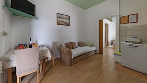A compact living space with a sofa, a dining table, a television mounted on a green shelf, and a washing machine. - Apartmani Mijatović (Lopar)