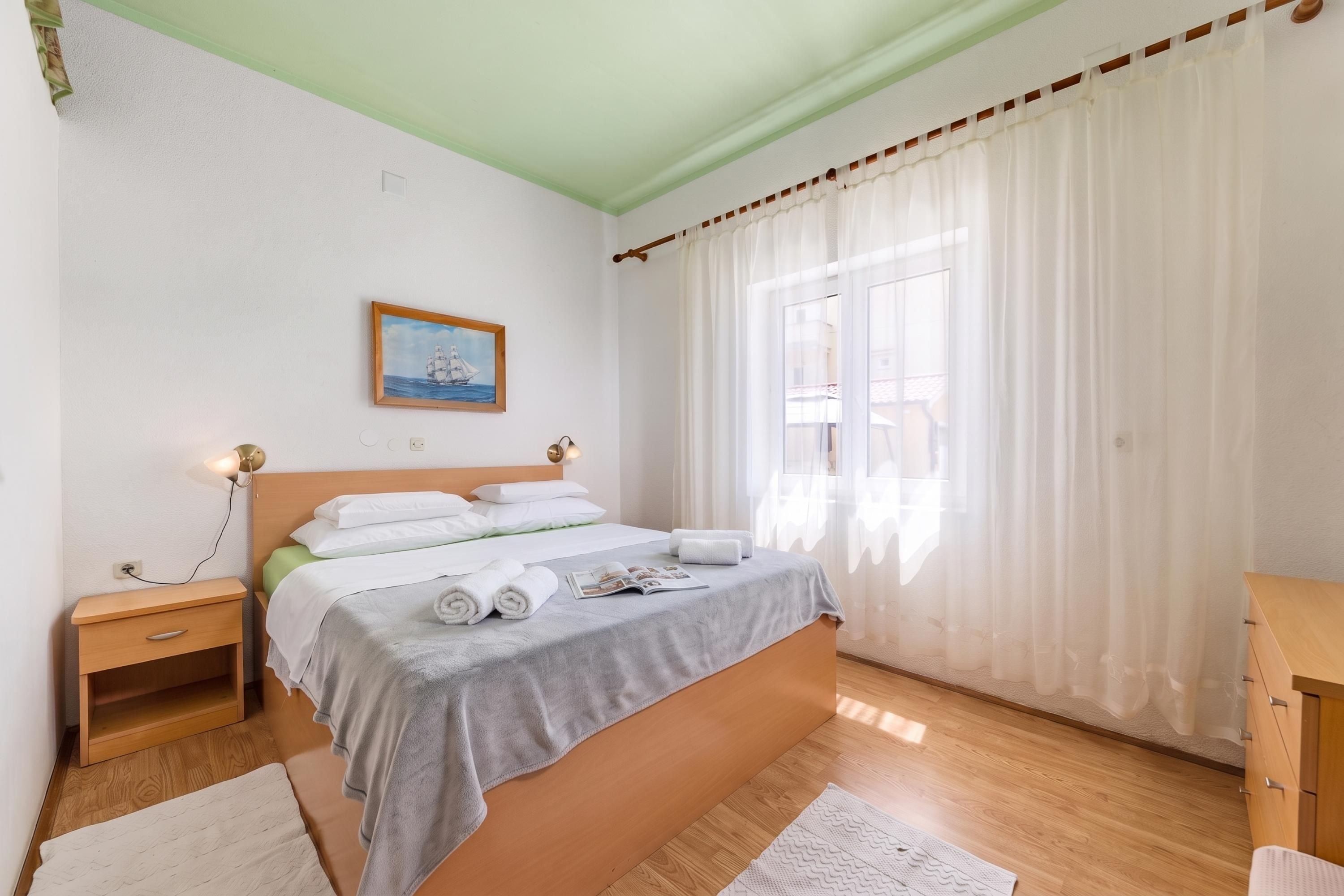 A bedroom with a bed, bedside tables, a nightstand, a window with curtains, and a painting on the wall.