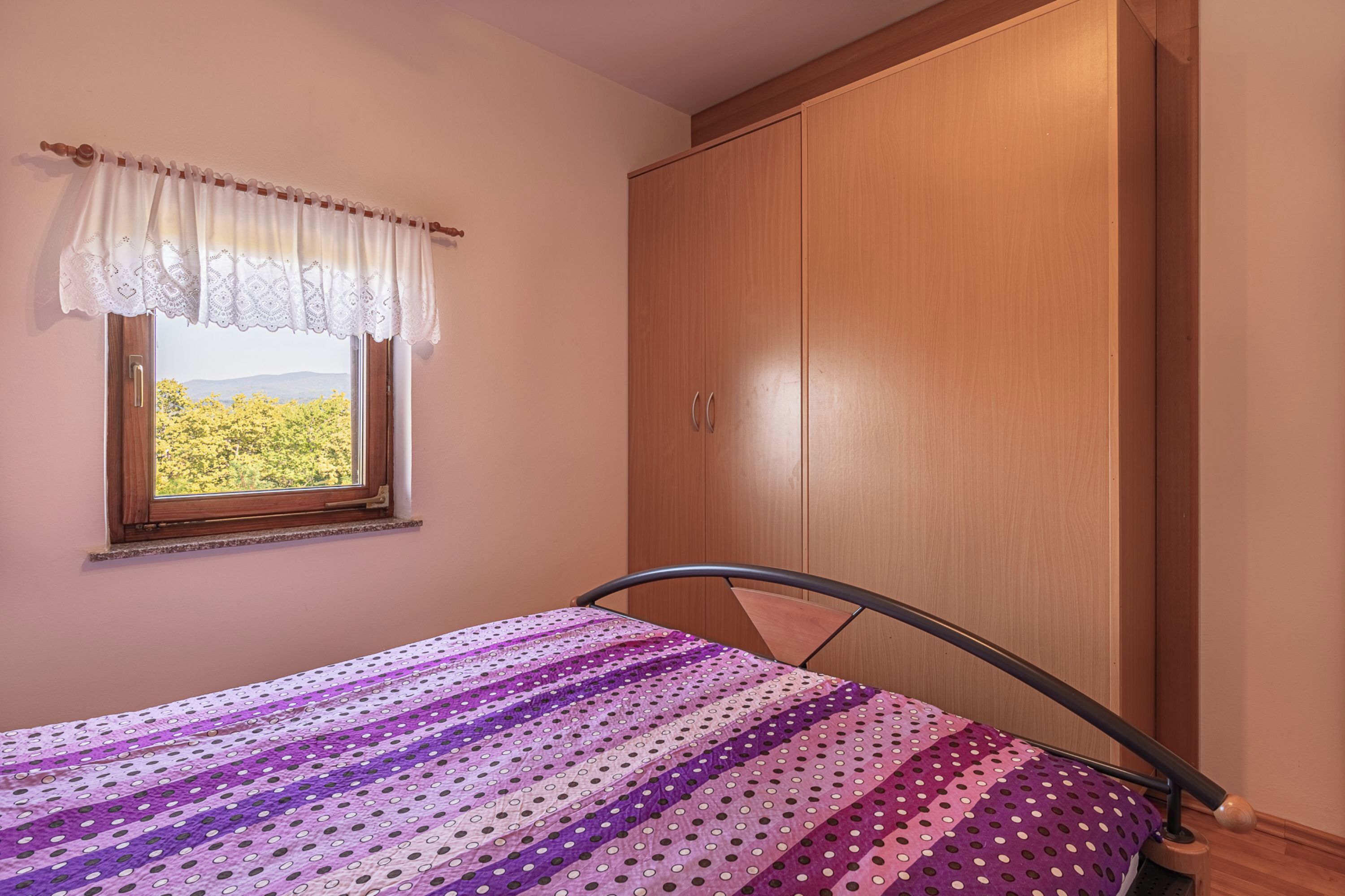 Een slaapkamer met een bed, een raam met uitzicht op groen en een houten garderobe.