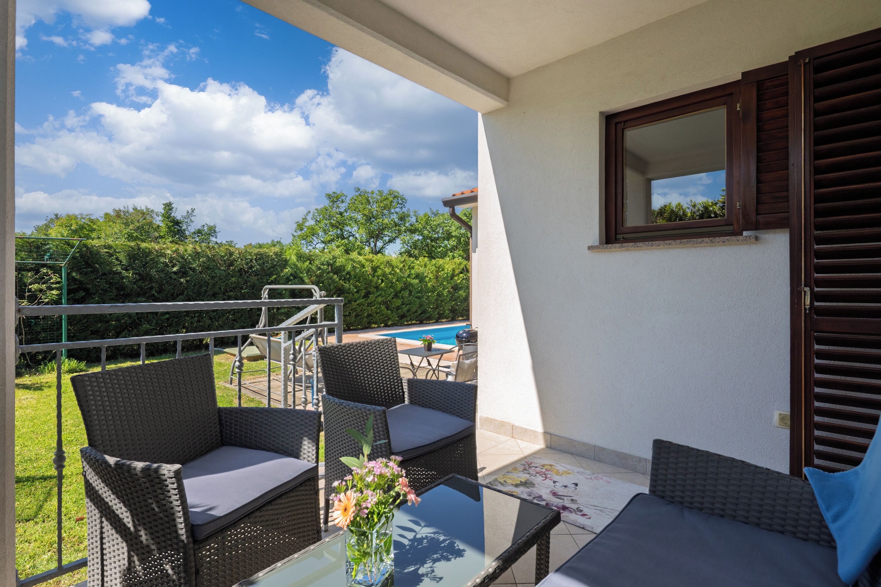 Een balkon met rieten meubilair, een glazen tafel en uitzicht op een zwembad en groen.