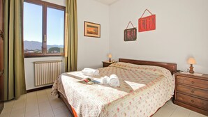 Un lit bien fait, recouvert d’une couvre-lit à motifs floraux, une table de chevet avec une lampe, une fenêtre avec des rideaux et un cadre photo accroché au mur.