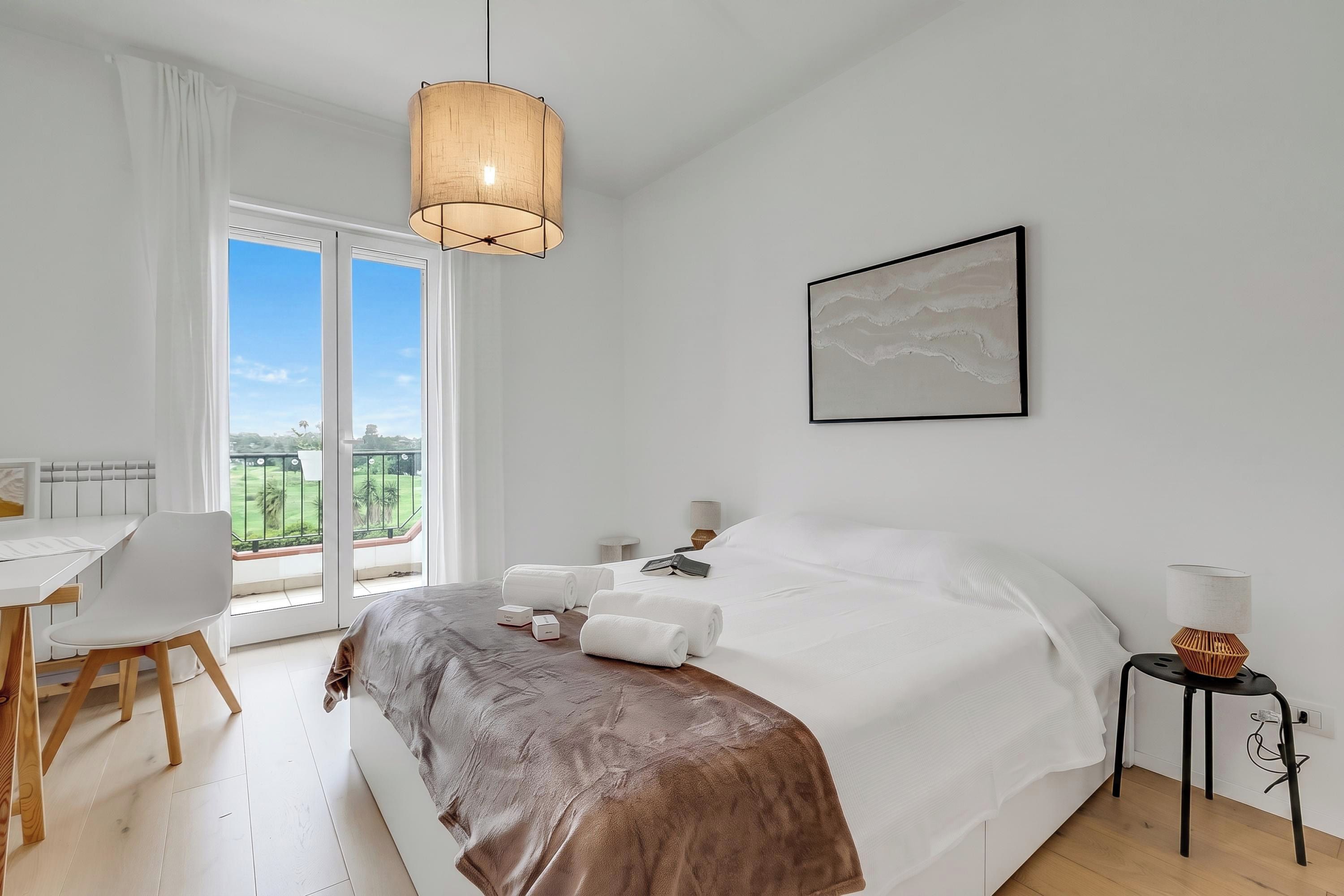 Habitación de hotel con cama, escritorio, silla, mesita de noche, lámpara y vista al exterior.