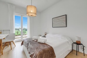 A hotel room with a bed, a desk, a chair, a bedside table, a lamp, and a view of the outdoors. - Sky View Apartment Monopoli (Monopoli)