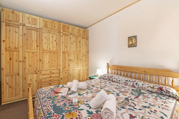 A bedroom with a wooden wardrobe, a bed with floral bedding, and a small bedside table. - Appartamento Quota 1800 for 4 + ski Storage (Claviere)
