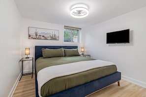 Un dormitorio con cama, lámparas de noche, televisor montado, ventana con persianas y ventilador de techo.