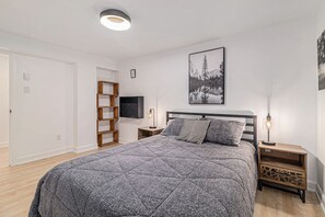 Ein Schlafzimmer mit einem Bett, einem an der Wand montierten Fernseher, einem Nachttisch mit Lampe und einer Holzregaleinheit.