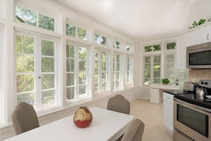 A kitchen with white cabinetry, a dining table with chairs, a microwave, an oven, and large windows with blinds.