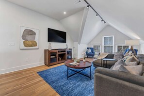 A spacious living room with a high ceiling, a flat-screen TV mounted on the wall, a wooden entertainment unit, a blue rug, and a grey sofa.