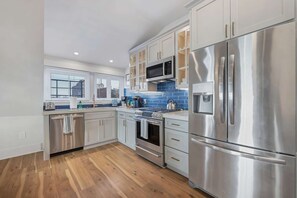 A modern kitchen with stainless steel appliances, wooden flooring, and a window with blinds.