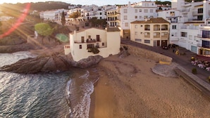 Un edificio frente al mar con balcón, junto a una playa de arena y un litoral rocoso.