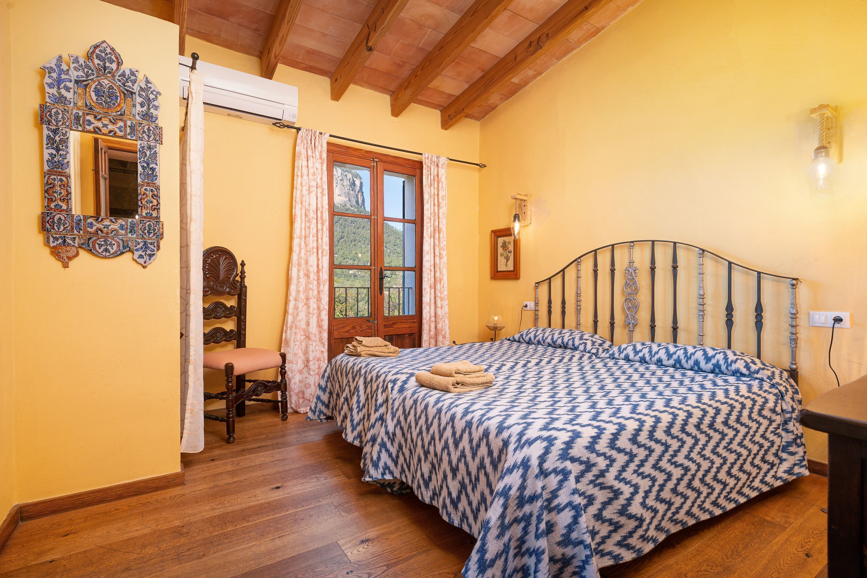 A bedroom with a wooden bed, a chair, a mirror, and a window with curtains.