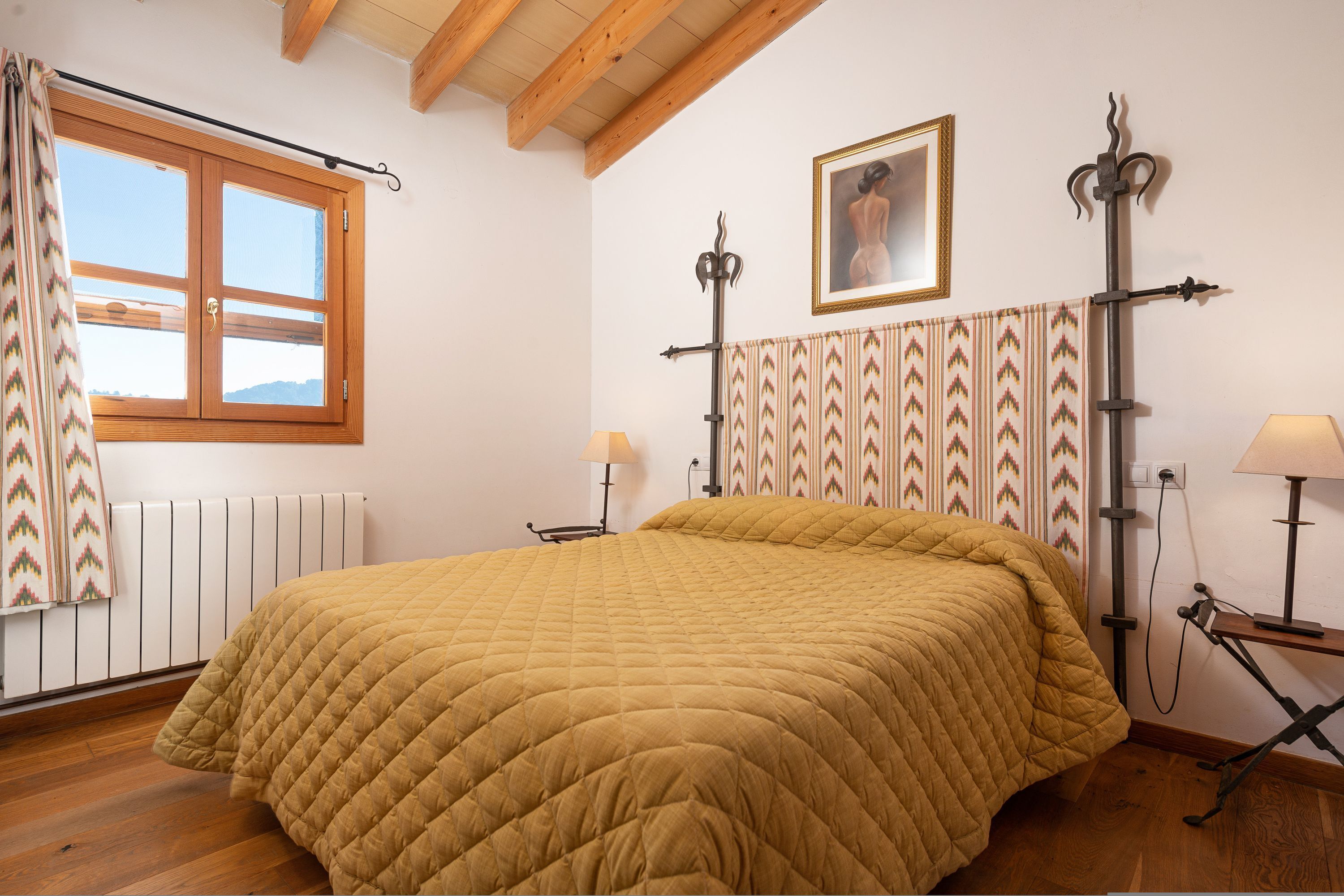 A bedroom with a large bed, a window with curtains, a radiator, a wall-mounted picture, and a decorative metal stand.