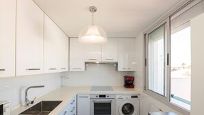 A modern kitchen with white cabinets, a stainless steel sink, a built-in oven, a washing machine, and a window providing a view of the outside. - Bajamar Deluxe in El Puerto de Santa Maria (El Puerto de Santa Maria)