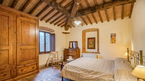 Un dormitorio con un armario de madera, una cama con colcha color crema, una silla, un aparador y un ventanal.