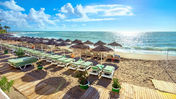 A beach with sun loungers and thatched umbrellas, a wooden deck, and a view of the ocean.