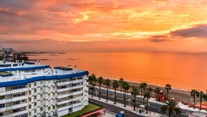 A coastal hotel with multiple floors, a parking area, and a view of the sea and sunset.