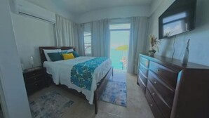 A bedroom with a bed, a dresser, a TV, and a view of the sea.