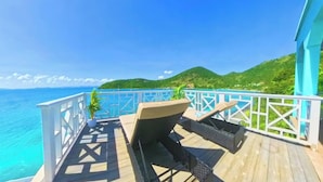 A wooden deck with a white railing overlooking a turquoise sea and green hills.