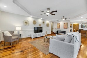 A modern living room with a flat-screen TV, a ceiling fan, and a dining area with a kitchen in the background. - Modern Historic 3 bdrm 2 bath (New Richmond)