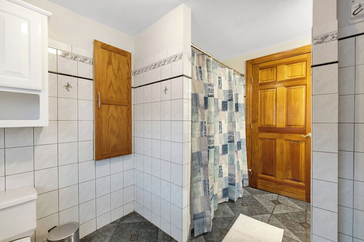 A bathroom with white tiled walls, a wooden door, a shower curtain, and a small trash bin.