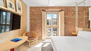 Quarto de hotel moderno com parede de tijolos, uma cama grande, uma cadeira de madeira, uma mesinha e uma janela ampla com vista.