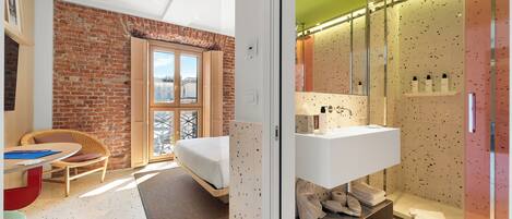 A modern room with a brick wall, a bed, a desk, and a bathroom with a white sink and shower.
