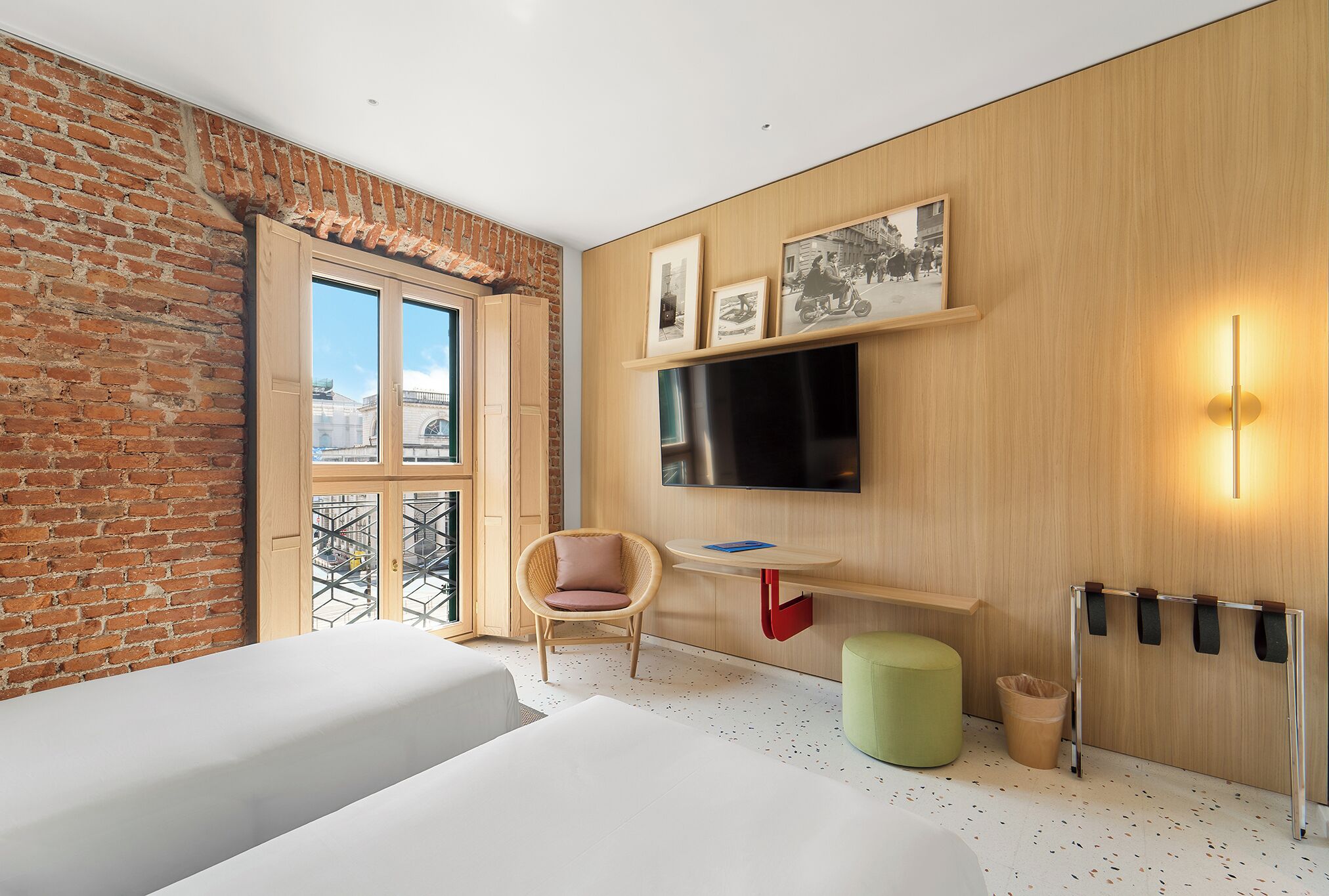 Ein modernes Hotelzimmer mit Backsteinwand, großem Fenster, Flachbildfernseher, Schreibtisch aus Holz, Korbstuhl, grünem Ottomanen und einem an der Wand befestigten Kleiderhaken.