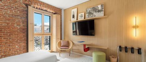 A modern hotel room with a brick wall, a large window, a flat-screen TV, a wooden desk, a wicker chair, a green ottoman, and a wall-mounted coat rack.