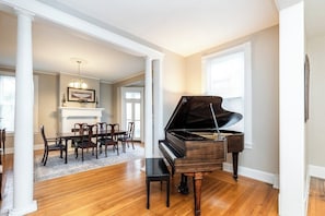 A grand piano in a spacious room with hardwood flooring, a dining area with chairs and a table, and a fireplace.