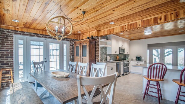 A spacious kitchen with wooden cabinets, a dining area with a long table and white chairs, and a view through large windows. - Cypress Chaos beautiful home with incredible marsh views in Willowbank (Georgetown)