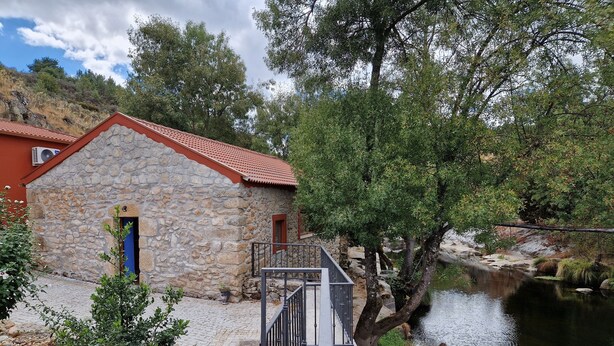 Un bâtiment en pierre avec un toit rouge, une porte bleue et un petit jardin parsemé de fleurs roses.