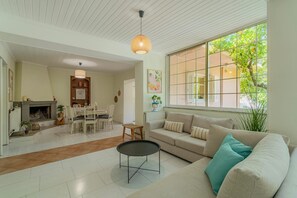 A living room with a sofa, a dining table, and chairs. There is a fireplace, a window with a view of greenery, and a hanging light fixture. - Joyful Secret Garden Residence Downtown, House (Chania)