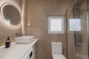 Un baño moderno con un espejo ovalado grande, un lavabo blanco, un inodoro y una ducha con mampara de vidrio.
