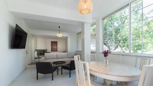 A modern living room with a sofa, dining table, and chairs. There is a TV mounted on the wall, and a window with a view of trees. - Joyful Secret Garden House by 8essentially (Chania)