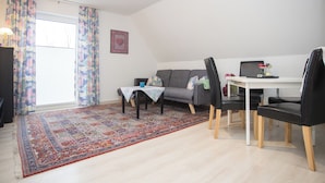 Un salon moderne avec un canapé gris, une table à manger avec des chaises, un tapis à motifs et une fenêtre avec des rideaux.
