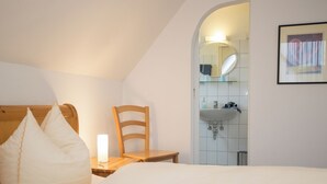 Een slaapkamer met een bed, kussens, een houten stoel, een nachtkastje met een lamp, een spiegel en een badkamer zichtbaar door een boog.