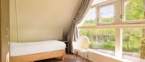Une chambre avec un lit simple, une chaise et de grandes fenêtres donnant sur un paysage verdoyant et une maison.