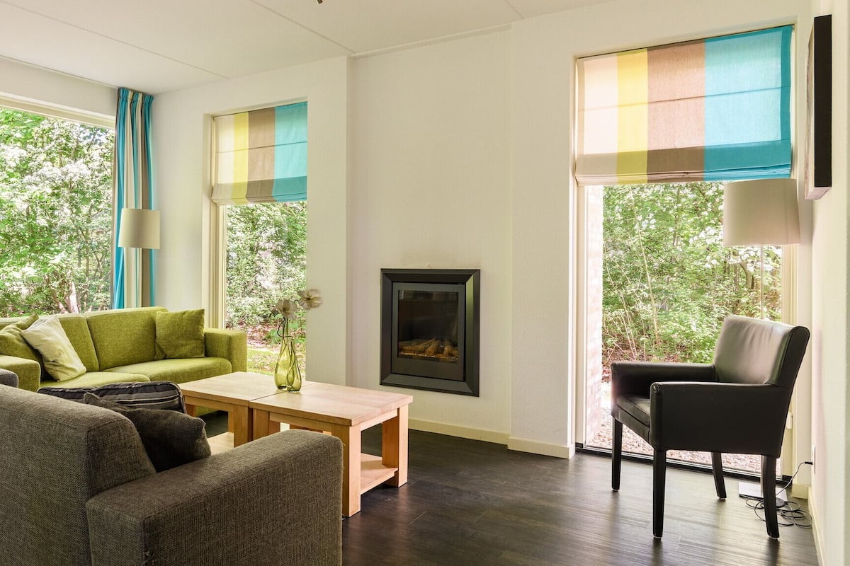 A living room with a green sofa, a grey sofa, a wooden coffee table, a fireplace, and large windows with curtains.