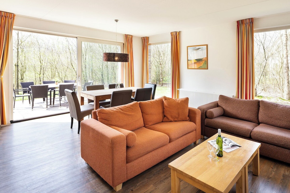 A modern living room with two sofas, a wooden coffee table, and a dining area with chairs and a table. Large windows provide a view of the outdoors.