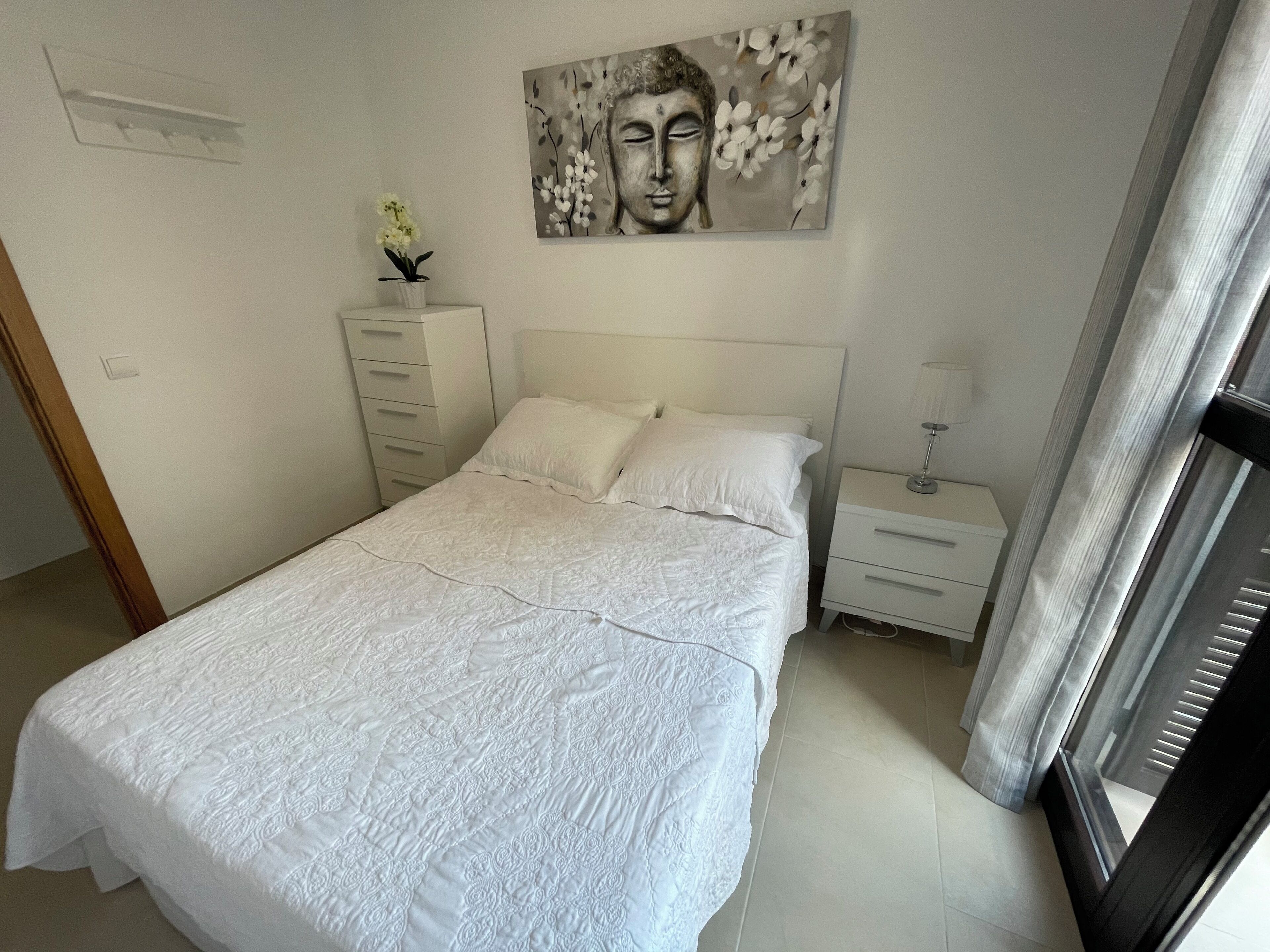 A bedroom with a large bed, a nightstand, a lamp, a wall-mounted artwork, and a window with curtains.